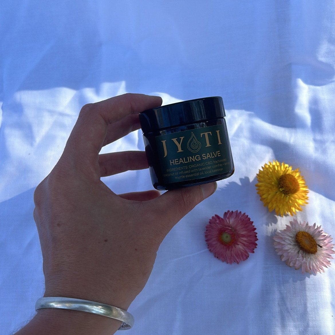JYOTI CBD Healing Salve displayed with organic ingredients on a white linen cloth with Australian native flowers